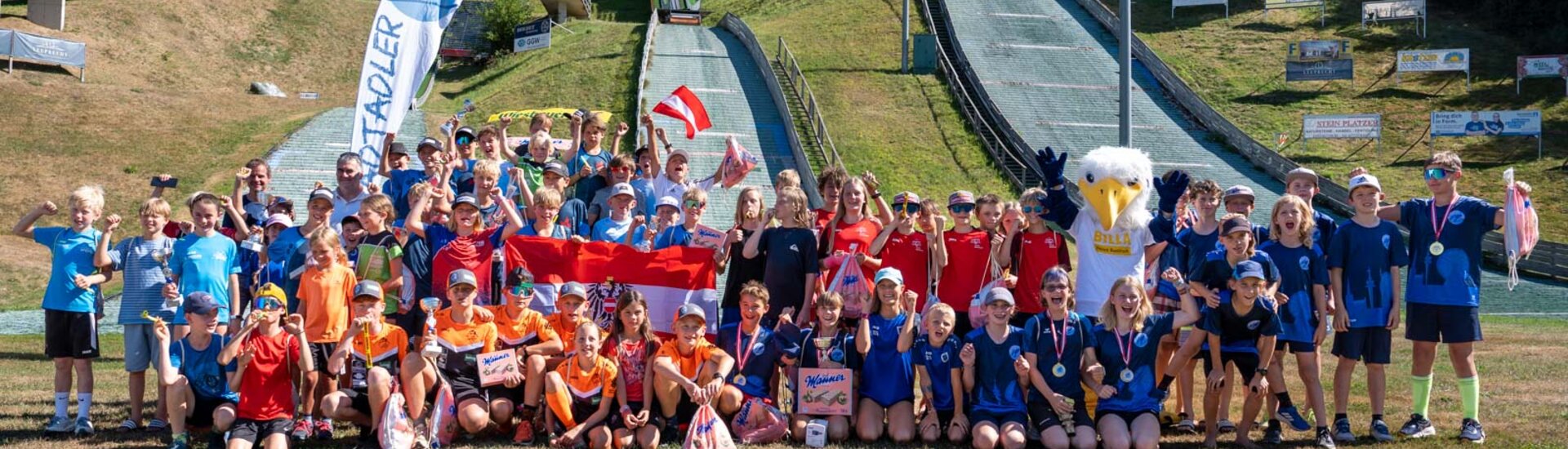 Alle teilnehmenden Kids der WienAIR 2026 stehen für Gruppenfoto zusammen im Schanzenauslauf, Sprungschanzen im Hintergrund zu sehen | © Albert Reiter