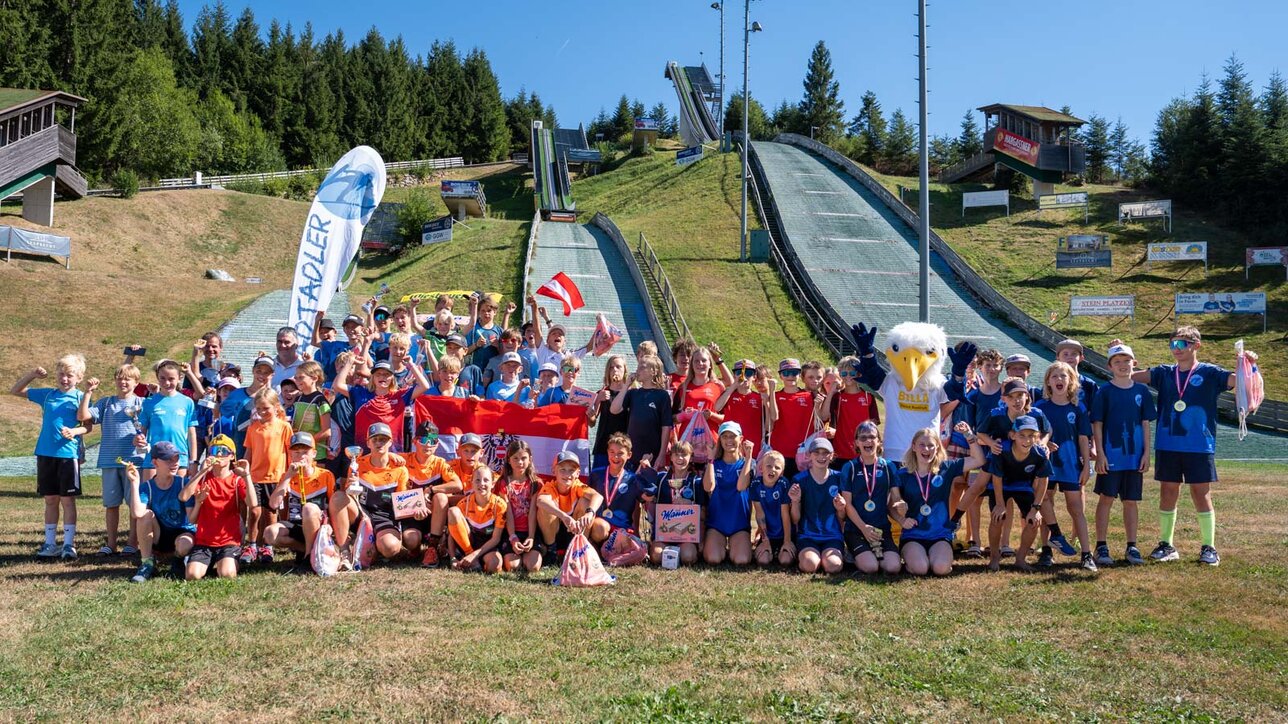 Alle teilnehmenden Kids der WienAIR 2026 stehen für Gruppenfoto zusammen im Schanzenauslauf, Sprungschanzen im Hintergrund zu sehen | © Albert Reiter