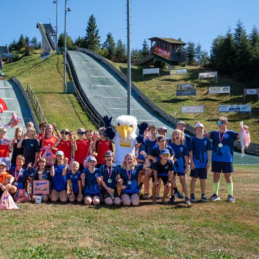Alle teilnehmenden Kids der WienAIR 2026 stehen für Gruppenfoto zusammen im Schanzenauslauf, Sprungschanzen im Hintergrund zu sehen | © Albert Reiter